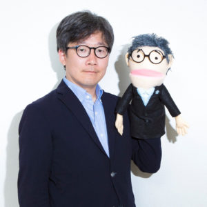 認知症分野のジャーナリストとして知られるコスガ聡一さん。コスガさんをイメージしたキャラクター人形「コッシ―」（右）と、全国の認知症カフェを訪問しレポートするなどの活動をおこなっている（コスガ聡一さん提供）