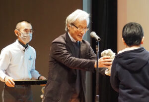 慶應義塾大学名誉教授で鶴見川流域ネットワーキング代表理事の岸由二さんが表彰状と景品の「バク」（鶴見川流域の形から生まれたキャラクター）のぬいぐるみを贈呈していた