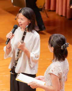 司会者からのインタビューに答える広報担当の丹藤さん。今回の演奏の実現に尽力してきた