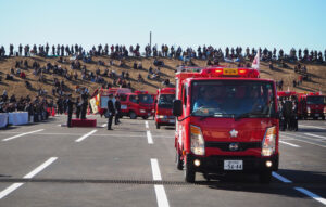消防団による車両分列行進の様子。観覧は駐車場内に設けられる観覧席のほか会場を見渡しやすい芝生エリアからも可能(2025年開催時)